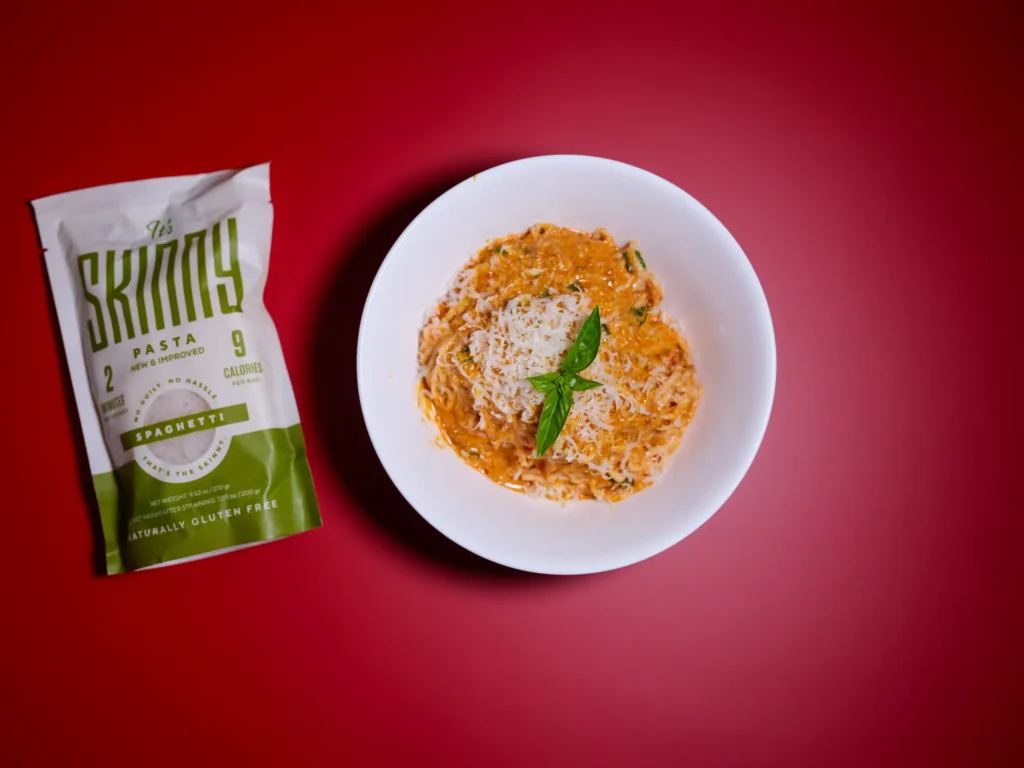 An overhead image of a white bowl of pasta on a red background, with a bag of It’s Skinny spaghetti noodles sitting on the surface next to the bowl. The bowl is filled with noodles coated in a creamy tomato sauce, topped with parmesan cheese and basil leaves.