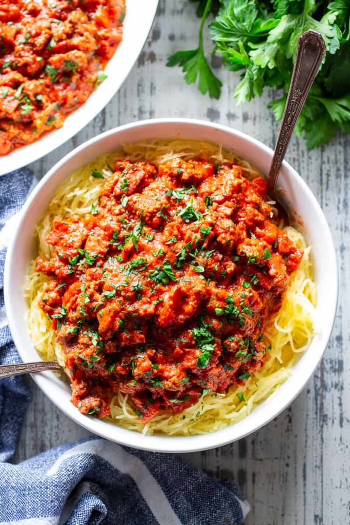 Red tomato Bolognese sauce sits on top of a bed of al dente spaghetti squash noodles in a white bowl, served with two spoons.