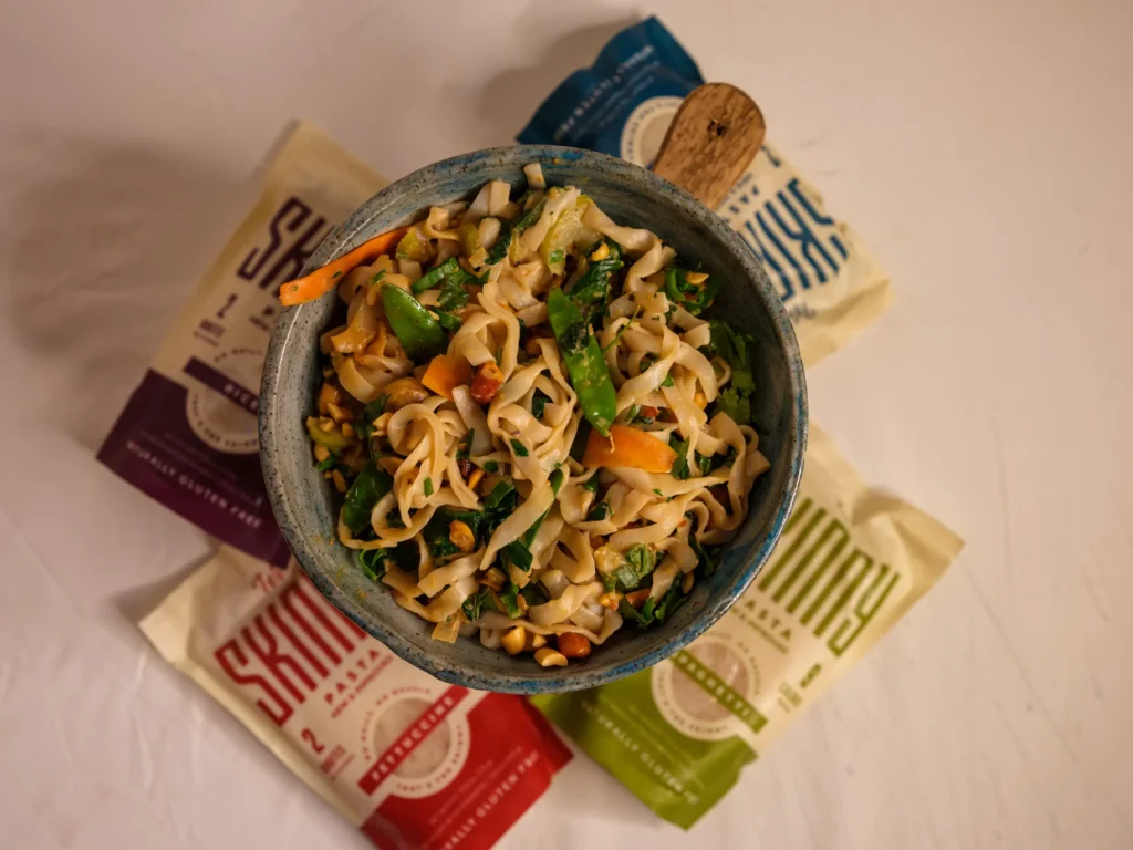 An overhead image of a full bowl of low-carb vegetable pad thai, made with It’s Skinny noodles, vegetables, and a tasty pad thai sauce. Four bags of It’s Skinny pasta surround the bowl.