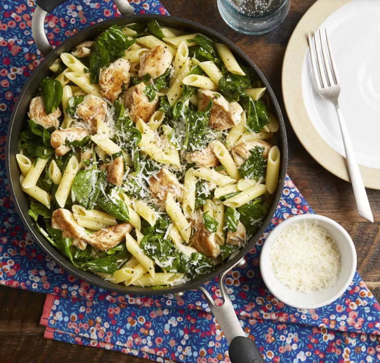 A spinach and penne pasta dish skillet sits next to a small bowl of parmesan cheese and an empty plate on a set table. 
